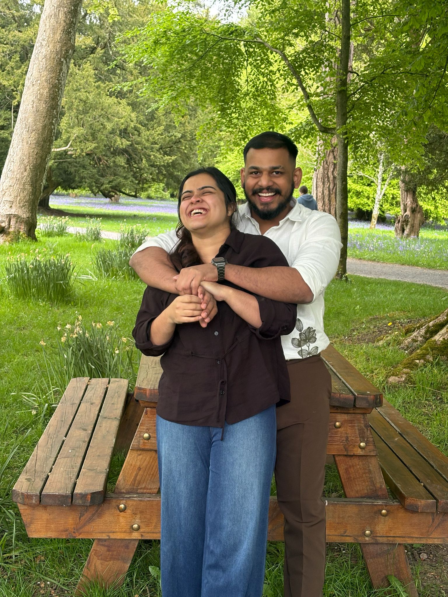 Leyo hugging Anju from behind as they laugh together beside a picnic table in the garden.